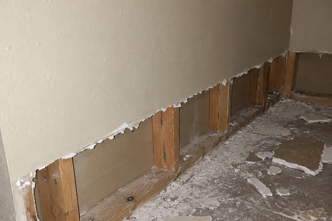 Drywall cut back above the floor along a wall, exposing studs, with cut pieces on the floor
